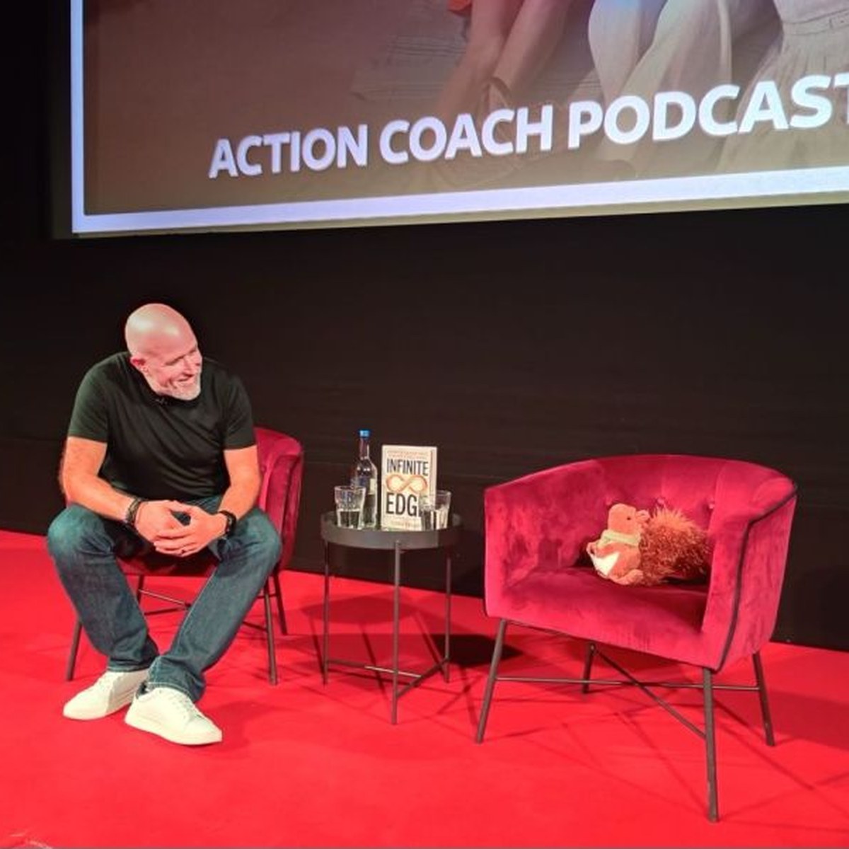 Neil smiling on a red velvet chair on stage, looking across at a toy squirrel sitting in the guest chair, with a podcast banner on the screen behind