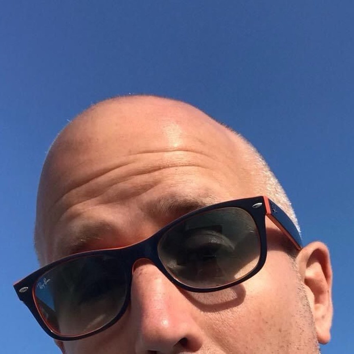 Neil in sunglasses peering up at the camera against a bright blue sky