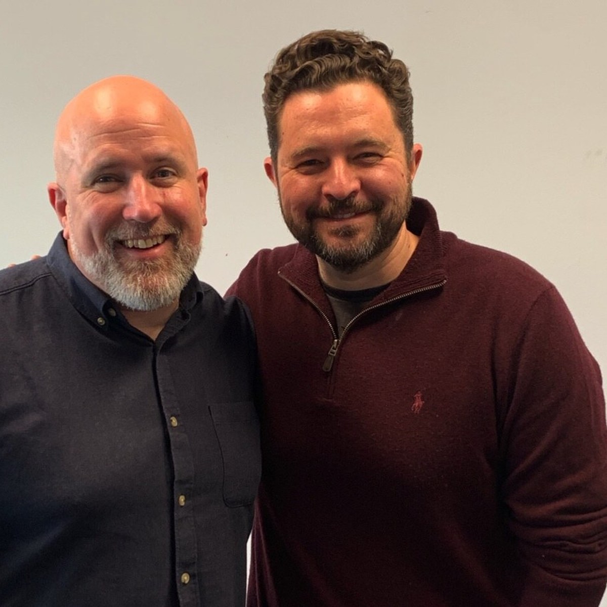 Neil Martin and Daniel Priestley standing side by side in a podcast studio with a brick wall and plants behind them