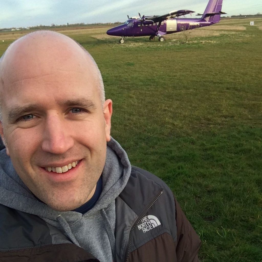 Neil Martin smiling at the airfield before his skydive, small plane on the grass behind