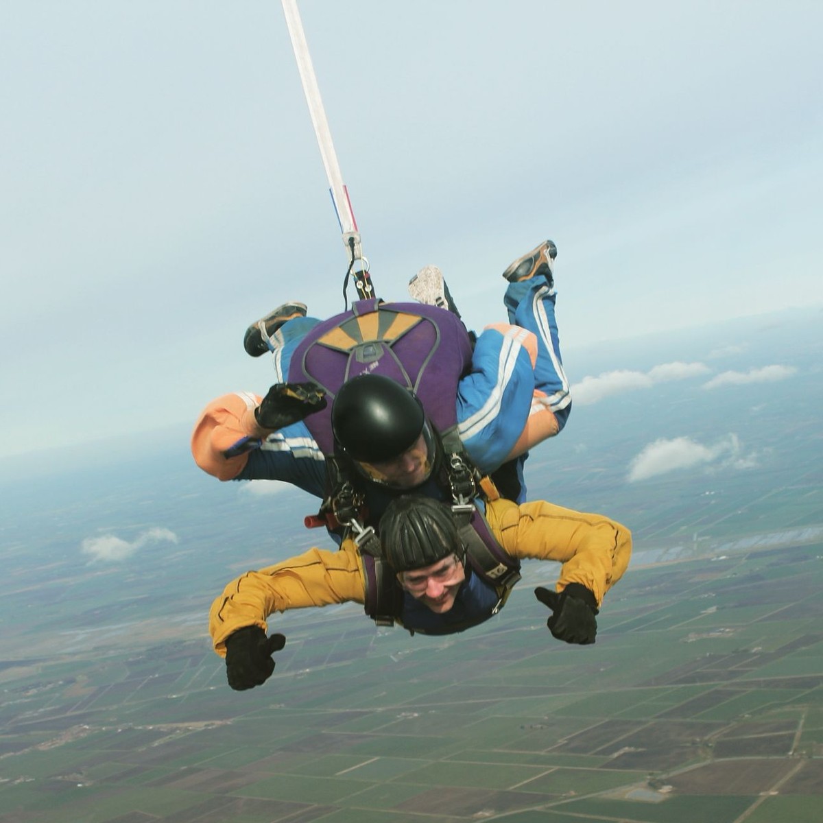 Neil Martin grinning in freefall on a tandem skydive, fields far below