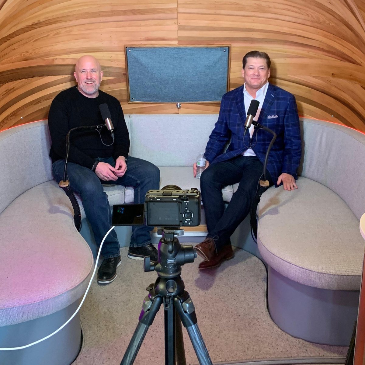 Neil Martin and Jeb Blount sitting in a curved wooden podcast pod at BizX 2026, with a camera on a tripod in front of them