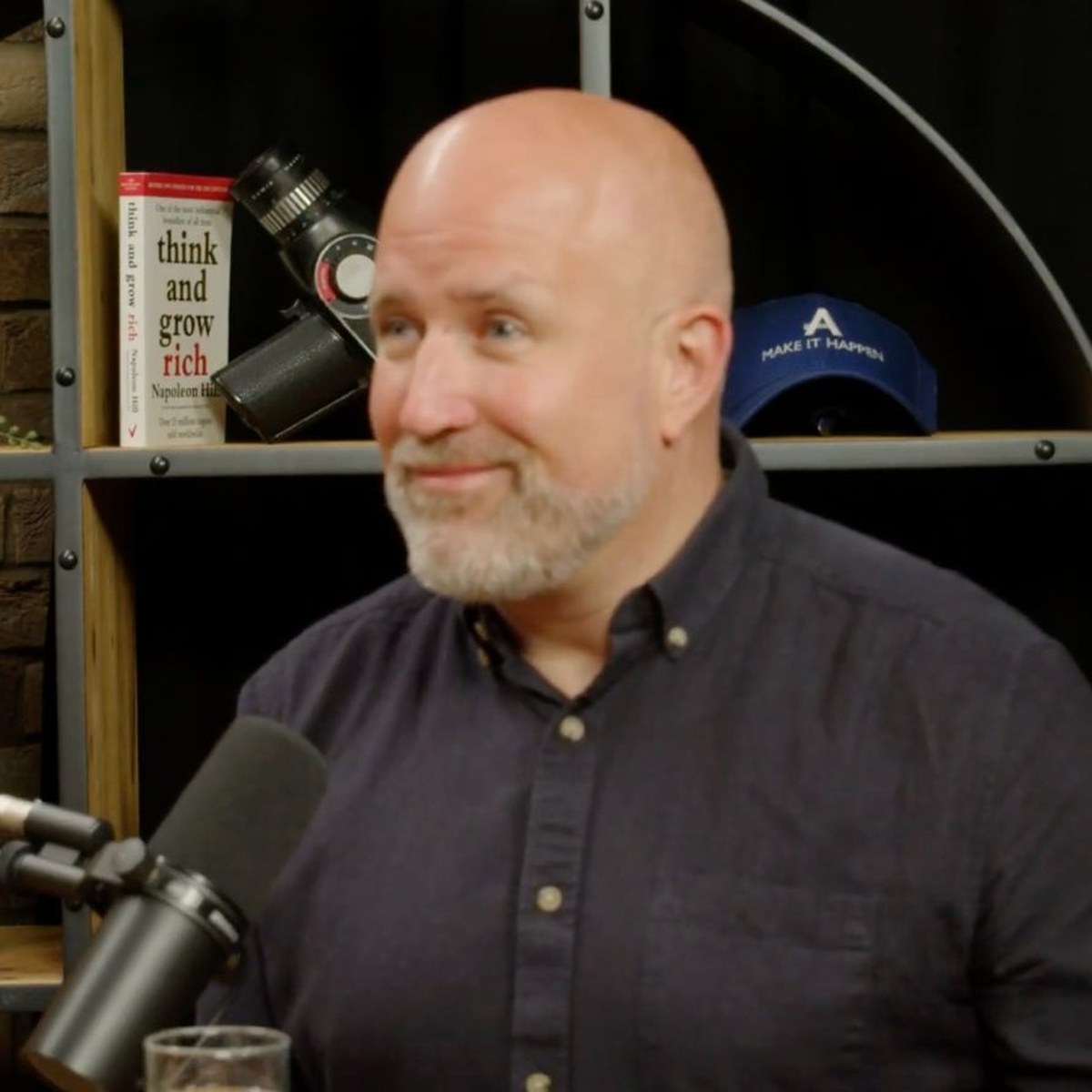 Neil Martin smiling at a microphone during an interview, with a copy of Think and Grow Rich on the shelf behind him