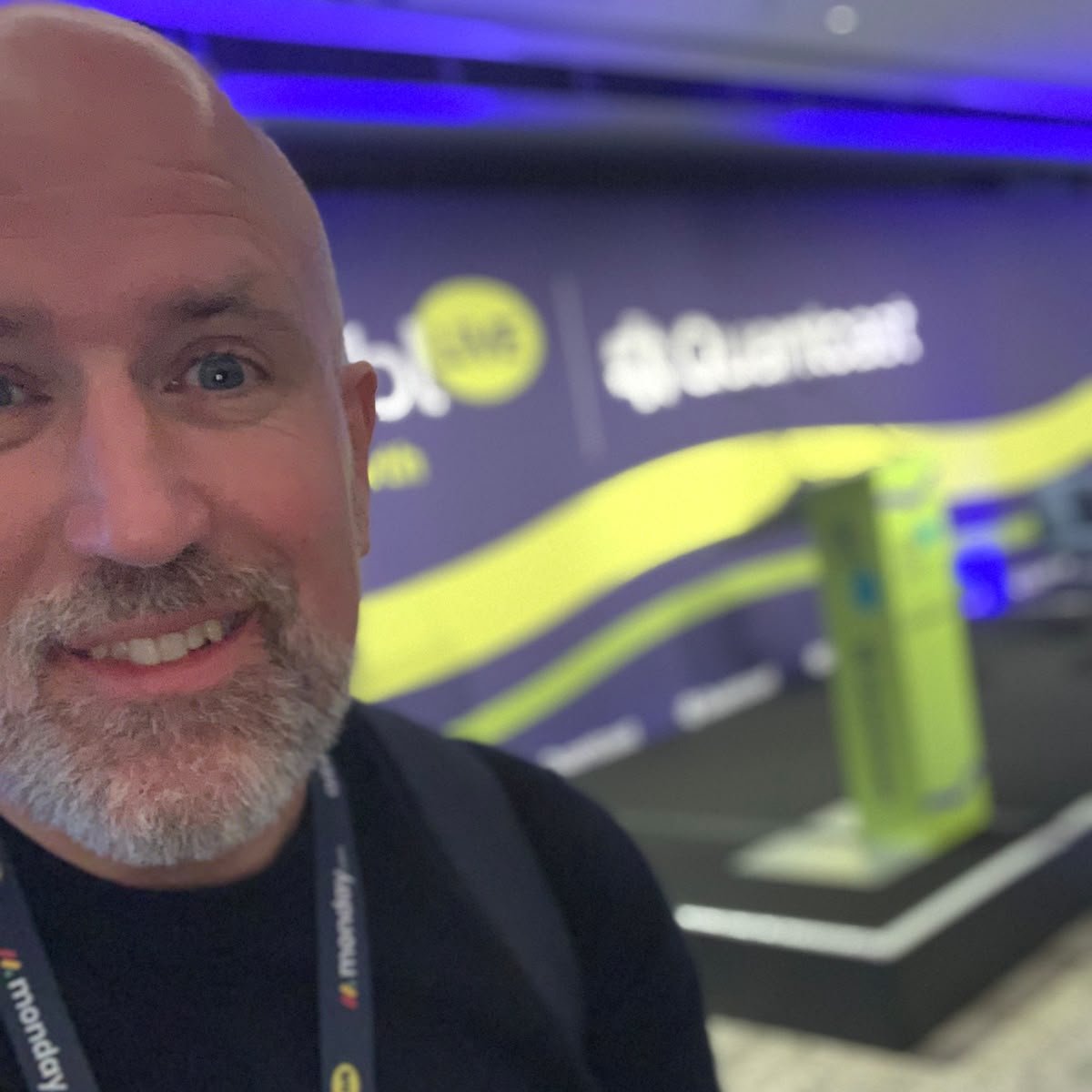 Neil Martin smiling in a selfie at aiblLIVE London, wearing his aibl lanyard, with the empty stage and branded backdrop behind him